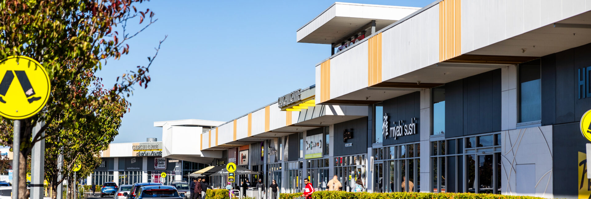 Exterior view of Churchill Centre Kilburn with cars parked and shoppers walking between stores on a sunny day.